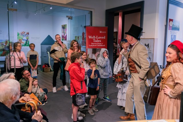 A man and woman in Victorian clothing stand in front of a crowd in a museum, and they are all stroking their chins as part of a performance.